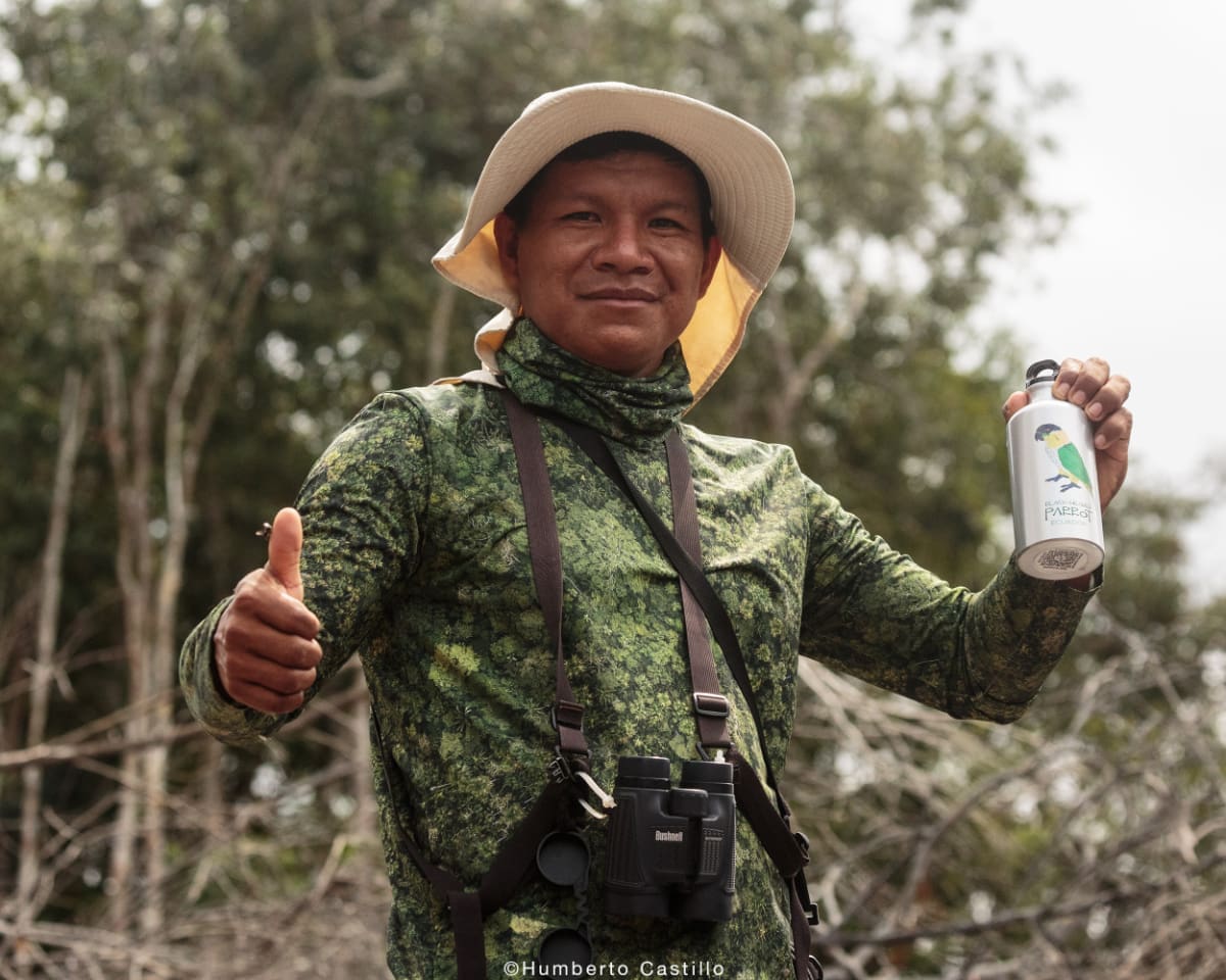 Kichwa man wearing birdwatching gear hold water bottle in left hand and thumbs up on his right