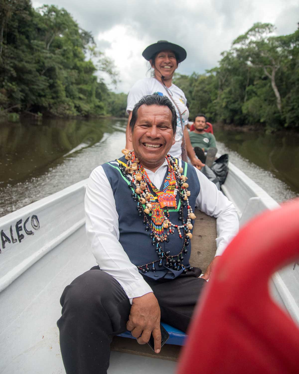 A Kichwa Shaman dressed in white shirt and blue vest wears necklaces and amulets while sitting in a motorized canoe